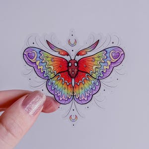May include: A rainbow-colored moth sticker with a heart on each wing. The moth is surrounded by stars and crescent moons.