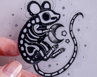 May include: A black and white illustration of a skeleton mouse holding a crescent moon. The mouse is surrounded by small stars.
