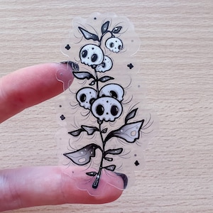 May include: A clear sticker featuring a black and white illustration of a plant with skull-shaped flowers. The sticker has a scalloped edge and is held by a finger. The design includes leaves and small, dark accents around the plant.
