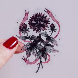 May include: A sticker featuring a dark purple flower with gray leaves and stems. Two pink, worm-like creatures with small faces encircle the flower. The sticker has a transparent background and a few small star-shaped accents.