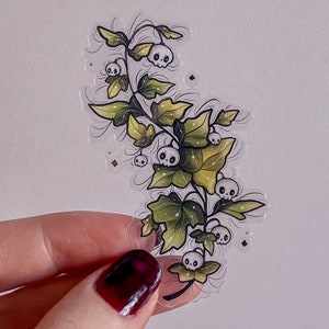 May include: A clear sticker featuring a whimsical illustration of a plant with green leaves and small white skulls. The leaves have a gradient of green and yellow. The sticker is held by a person's hand.