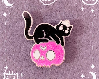 Può includere: Gatto nero con una corona seduto su un teschio rosa glitter con occhi bianchi e una mezzaluna sulla parte superiore.