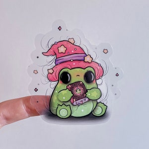 Puede incluir: Una rana verde con un sombrero de bruja rosa y morado sostiene un libro que dice "Frog Spells". La rana está rodeada de un fondo blanco con estrellas rosas.