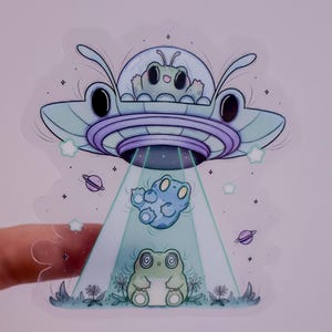 May include: A whimsical sticker featuring a cartoon UFO with a light blue and purple color scheme. The UFO is abducting a blue frog, while another frog with spiral eyes is on the ground. Stars and planets are in the background.