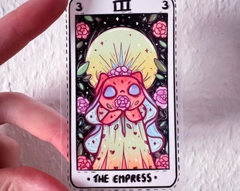 Puede incluir: Una pegatina transparente con una colorida ilustración de una figura felina sosteniendo una rosa, con una gran luna de fondo. La pegatina es una carta del tarot, etiquetada como "THE EMPRESS" y "III". El diseño incluye rosas rosas y hojas verdes.