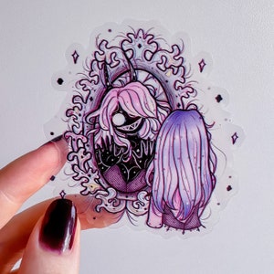 Op de afbeelding: Een sticker met een zwart-witte illustratie van een vrouw met hoorns en een sinistere glimlach die in een spiegel kijkt. De vrouw in de spiegel heeft roze haar en draagt een zwarte jurk met een visnetpatroon. De sticker heeft een paars en roze kleurenschema en is omgeven door een decoratief frame.