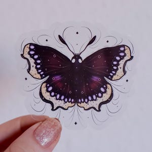 Könnte beinhalten: Ein gestanzter Aufkleber mit einem dunkelvioletten und schwarzen Schmetterling mit cremefarbenen Flügelrändern. Der Schmetterling hat weiße kreisförmige Markierungen auf dem Körper, hellviolette Punkte an den Flügelrändern und feine schwarze Linien.