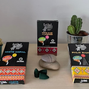 May include: Three boxes of incense cones with different scents: White Copal, White Sage + Lavender, and Palo Santo. The boxes are black with white text and feature a Native American dreamcatcher design. The boxes are sitting on a wooden table with two small green cacti in white pots and a stack of grey stones.