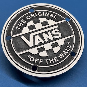 Peut inclure: Un emblème métallique circulaire avec une finition noire et argentée et un motif en damier. Le texte indique THE ORIGINAL VANS OFF THE WALL. Le design comprend quatre petits trous circulaires positionnés sur le bord extérieur pour la fixation.