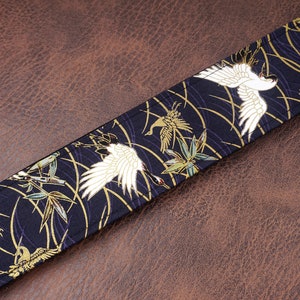 May include: A fabric strap featuring a dark navy blue background with a repeating pattern of white cranes, green bamboo leaves, and thin gold curved lines. The strap has visible stitching along the edges and a flat, rectangular construction.