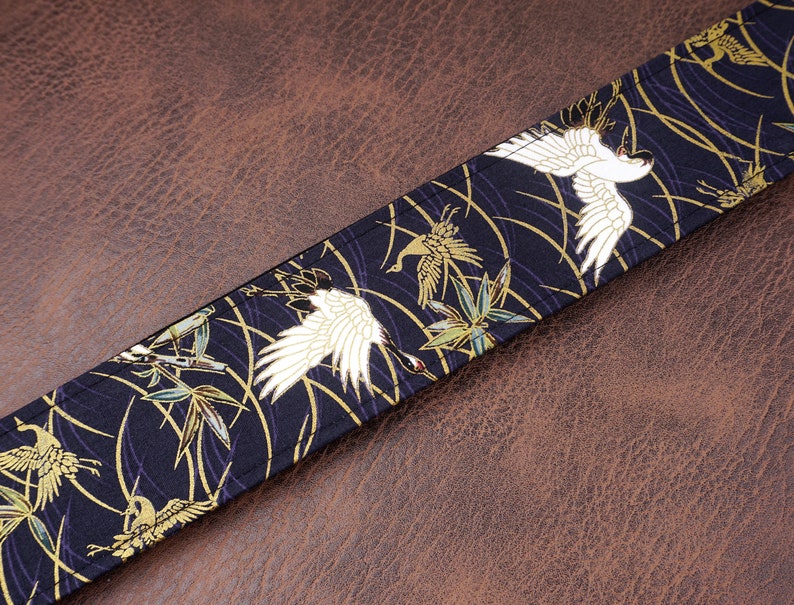 May include: A fabric strap featuring a dark navy blue background with a repeating pattern of white cranes, green bamboo leaves, and thin gold curved lines. The strap has visible stitching along the edges and a flat, rectangular construction.