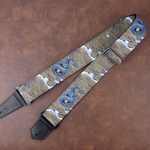 Op de afbeelding: Een gitaarband met een blauw en gouden draken- en golfpatroon. De band heeft zwarte leren uiteinden met een metalen gesp.