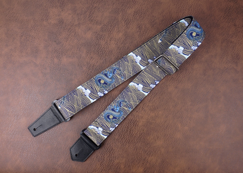 Op de afbeelding: Een gitaarband met een blauw en gouden draken- en golfpatroon. De band heeft zwarte leren uiteinden met een metalen gesp.