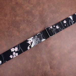 Peut inclure: Sangle de guitare noire avec un motif floral blanc et rose repr&eacute;sentant des carpes ko&iuml;. La sangle est dot&eacute;e d'une boucle argent&eacute;e r&eacute;glable.