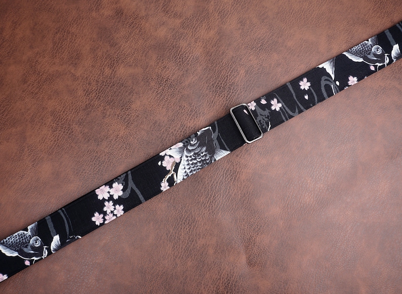 Peut inclure: Sangle de guitare noire avec un motif floral blanc et rose repr&eacute;sentant des carpes ko&iuml;. La sangle est dot&eacute;e d'une boucle argent&eacute;e r&eacute;glable.