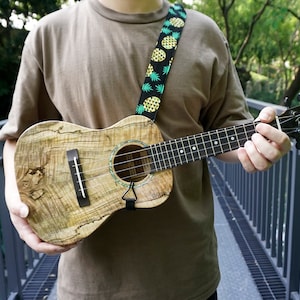 Może przedstawiać: Osoba grająca na brązowym ukulele z czarnym i zielonym paskiem w ananasowy wzór. Ukulele ma czarną główkę z napisem "Ranier".