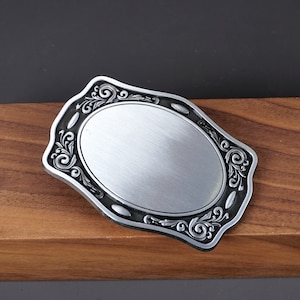 Pode incluir: Fivela de cinto prateada com um espaço em branco oval no centro e um design ornamentado e rolado ao redor da borda.