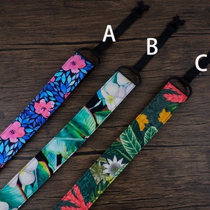 May include: Three camera straps with different floral patterns. The straps are black with adjustable lengths. The first strap has a blue background with pink flowers. The second strap has a white background with green leaves and pink flowers. The third strap has a green background with red, yellow, and white flowers.