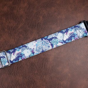 May include: A guitar strap featuring a dark blue background with a repeating pattern of white and light blue chrysanthemums and koi fish. The strap includes a silver metal adjustment buckle and a black leather end tab with a single hole for attachment.