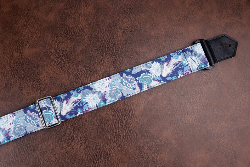 May include: A guitar strap featuring a dark blue background with a repeating pattern of white and light blue chrysanthemums and koi fish. The strap includes a silver metal adjustment buckle and a black leather end tab with a single hole for attachment.