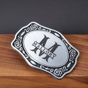 May include: A rectangular silver-toned metal belt buckle with rounded, scalloped edges featuring an ornate scrollwork border. The center has a brushed silver finish with a large black letter M and the name MALLORY printed horizontally across it.