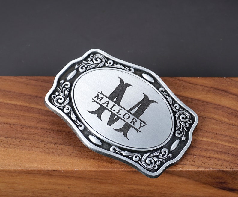 May include: A rectangular silver-toned metal belt buckle with rounded, scalloped edges featuring an ornate scrollwork border. The center has a brushed silver finish with a large black letter M and the name MALLORY printed horizontally across it.