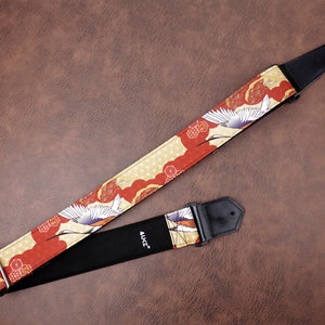 May include: A red and gold guitar strap with a white crane design. The strap is made of fabric and has a black leather end with the word "4UGC" printed on it.
