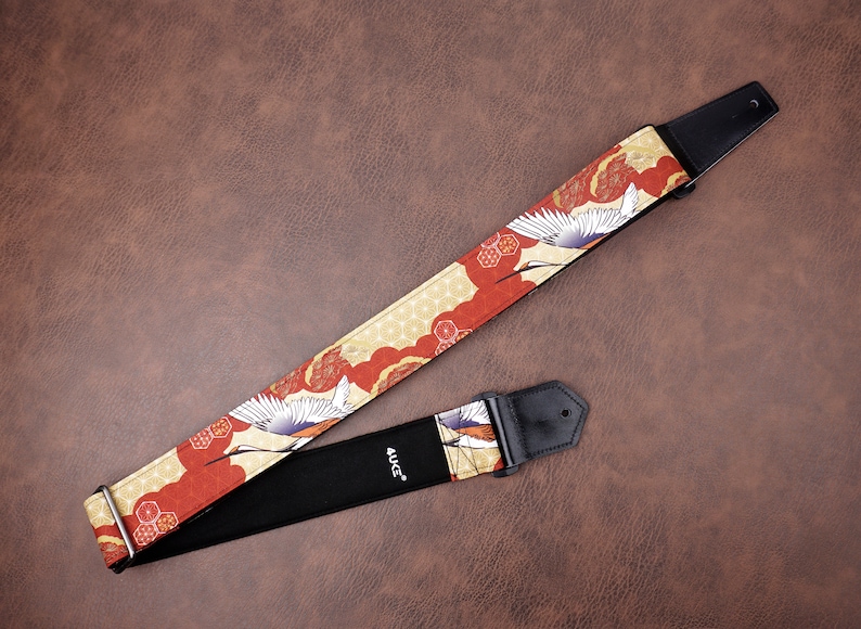 May include: A red and gold guitar strap with a white crane design. The strap is made of fabric and has a black leather end with the word "4UGC" printed on it.