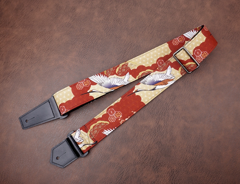 May include: A red and gold guitar strap with a Japanese crane pattern. The strap has black leather ends.