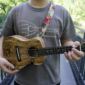 Może przedstawiać: Osoba grająca na brązowym i czarnym ukulele z kwiatowym paskiem do ukulele. Ukulele ma czarną główkę z napisem "Renovo".