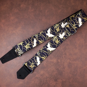May include: A guitar strap featuring a dark navy blue fabric with a repeating pattern of white cranes and gold bamboo stalks. The strap includes black leather end tabs with pre-cut holes and a silver-toned metal adjustable sliding buckle.
