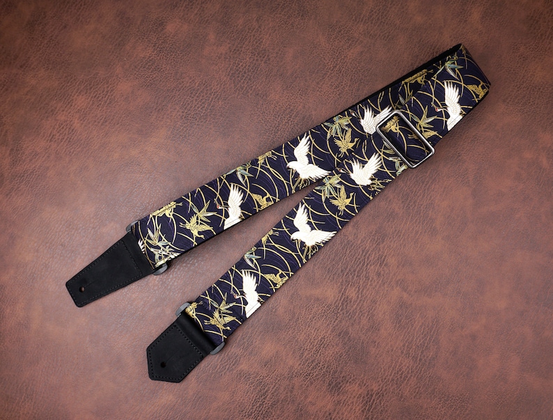 May include: A guitar strap featuring a dark navy blue fabric with a repeating pattern of white cranes and gold bamboo stalks. The strap includes black leather end tabs with pre-cut holes and a silver-toned metal adjustable sliding buckle.