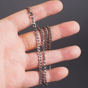 Puede incluir: Un collar de cadena de eslabones de color plateado. La cadena está hecha de pequeños eslabones entrelazados y tiene un cierre de mosquetón. El collar se muestra sobre un fondo oscuro.