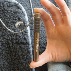 May include: A bronze-coloured pen-shaped pendant with a textured grip and a silver chain. The pendant has the engraved name "Luca Balboni" in a cursive font. The pen is held in a hand against a grey background.