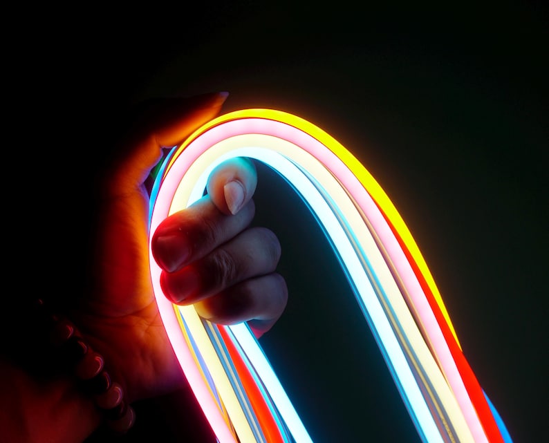May include: A hand holds a flexible, multi-colored neon light strip against a dark background. The strip features vibrant colors including red, orange, yellow, pink, and blue. The neon lights are curved, creating an arc shape.
