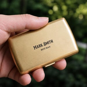 May include: A gold-colored metal business card holder. The text "MARK SMITH BEST MAN" is printed in black on the front of the holder. The holder is rectangular with rounded edges and is held in a person's hand.