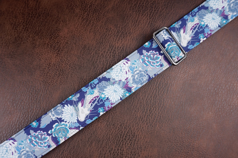 May include: A fabric strap featuring a repeating pattern of white and light blue koi fish interspersed with blue, white, and purple floral motifs. The strap includes a rectangular silver-toned metal slider for length adjustment and stitching.