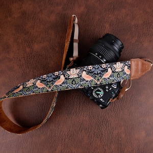 May include: A camera strap featuring a repeating pattern of light orange birds and floral motifs on a dark background, accented with tan leather ends and adjustable beige webbing straps, attached to a black digital camera body.