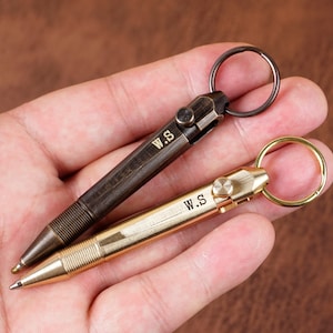 May include: Two keychains with a pen tip. One keychain is brass with a black pen tip and the other is brass with a gold pen tip. Both keychains have the initials "W.S." engraved on them.