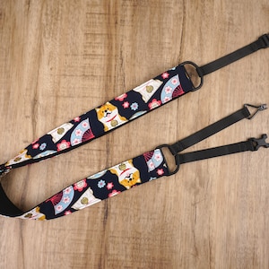 Puede incluir: Una correa de cámara negra con un patrón colorido de perros Shiba Inu y abanicos japoneses. La correa tiene longitud ajustable y cuenta con una hebilla de liberación rápida.