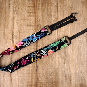 Puede incluir: Una correa de cámara negra con un estampado floral colorido. La correa tiene longitud ajustable y presenta un anillo metálico negro y dos clips de plástico negro.