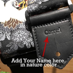 May include: A guitar strap featuring a black fabric section with a white crane and gray floral pattern, connected to a black leather end piece. The leather tab is embossed with the name Carrel and includes a circular hole for attachment.