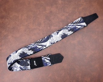 Peut inclure: Une sangle de guitare noire et blanche avec un motif de vagues bleues. La sangle a une extrémité en cuir noir avec le mot "AUKE" imprimé dessus.