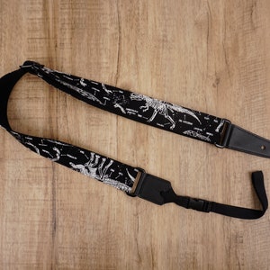 Puede incluir: Correa de guitarra negra con estampado de esqueleto de dinosaurio blanco. La correa tiene un extremo de cuero negro con una hebilla de metal.