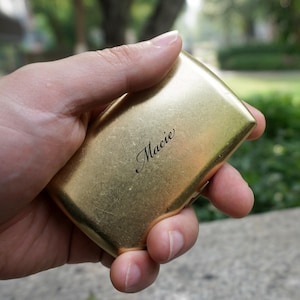 May include: A gold-colored, rectangular compact held in a hand. The compact has a slightly rounded shape and a shiny finish. The name "Macie" is elegantly inscribed on the surface in a cursive font. The background is blurred, suggesting an outdoor setting.