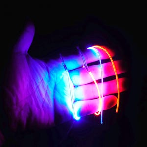 Op de afbeelding: Een hand verlicht met neonlichten in tinten blauw, rood en oranje. De lichten lijken aan de vingers bevestigd te zijn, waardoor een gloeiend effect ontstaat tegen een donkere achtergrond. Het algehele effect is futuristisch en levendig.