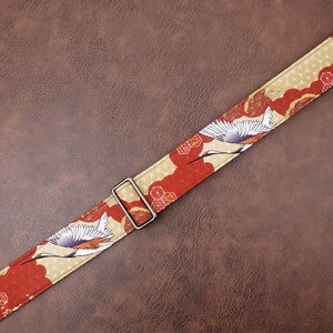 May include: A red and gold guitar strap with a Japanese crane design. The strap has a silver buckle.