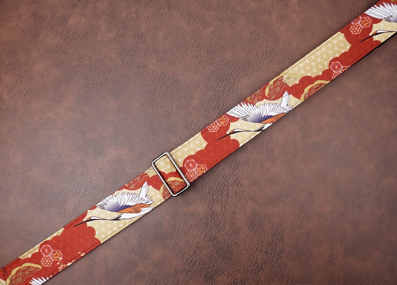 May include: A red and gold guitar strap with a Japanese crane design. The strap has a silver buckle.