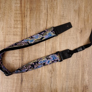 Puede incluir: Una correa de guitarra negra con un patrón floral azul y dorado. La correa tiene una hebilla ajustable negra y un extremo de cuero negro.