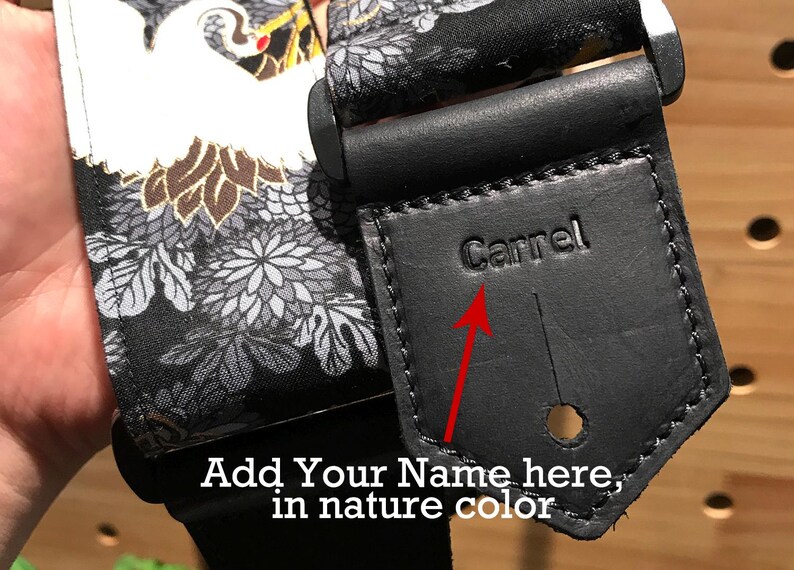 May include: Black guitar strap with a white and gold floral pattern. The strap has a black leather patch with the word "Carrel" embossed on it. The patch has a space for adding a name in nature color.